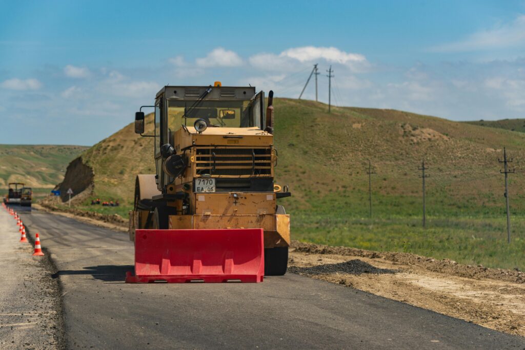 A road roller compacts asphalt on a rural road with green hills in the background; heavy machinery in action.